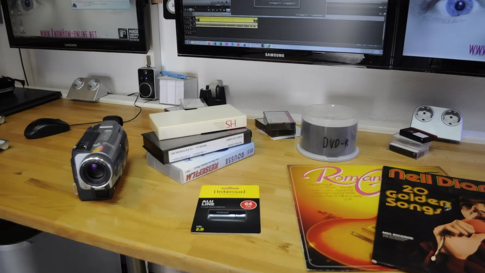 Arbeitstisch im Labor von KNOW HOW: Camcorder, VHS-Kassetten, eine Spindel DVD-Rohlinge, ein USB-Stick und zwei Schallplatten, dahinter Monitore.