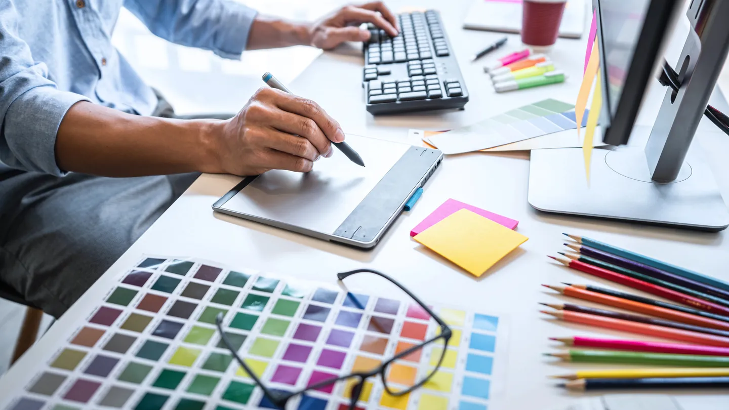 Grafikarbeitsplatz: Eine Hand zeichnet mit dem Stift auf einem Grafiktablett, davor Farbmuster, Buntstifte und eine Brille.