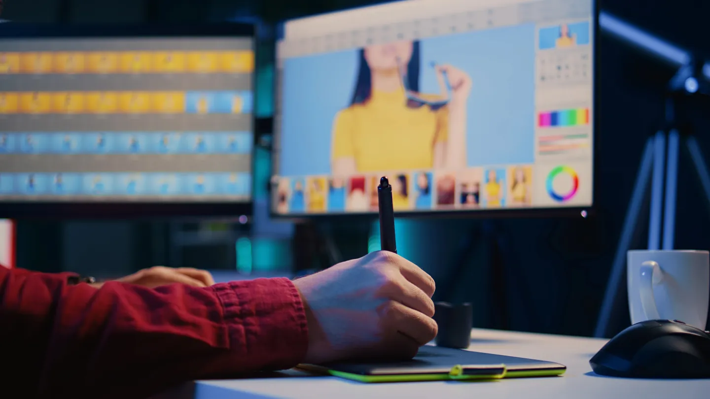 Eine Hand retuschiert mit Stift und Grafiktablett ein Porträtfoto auf dem Monitor.