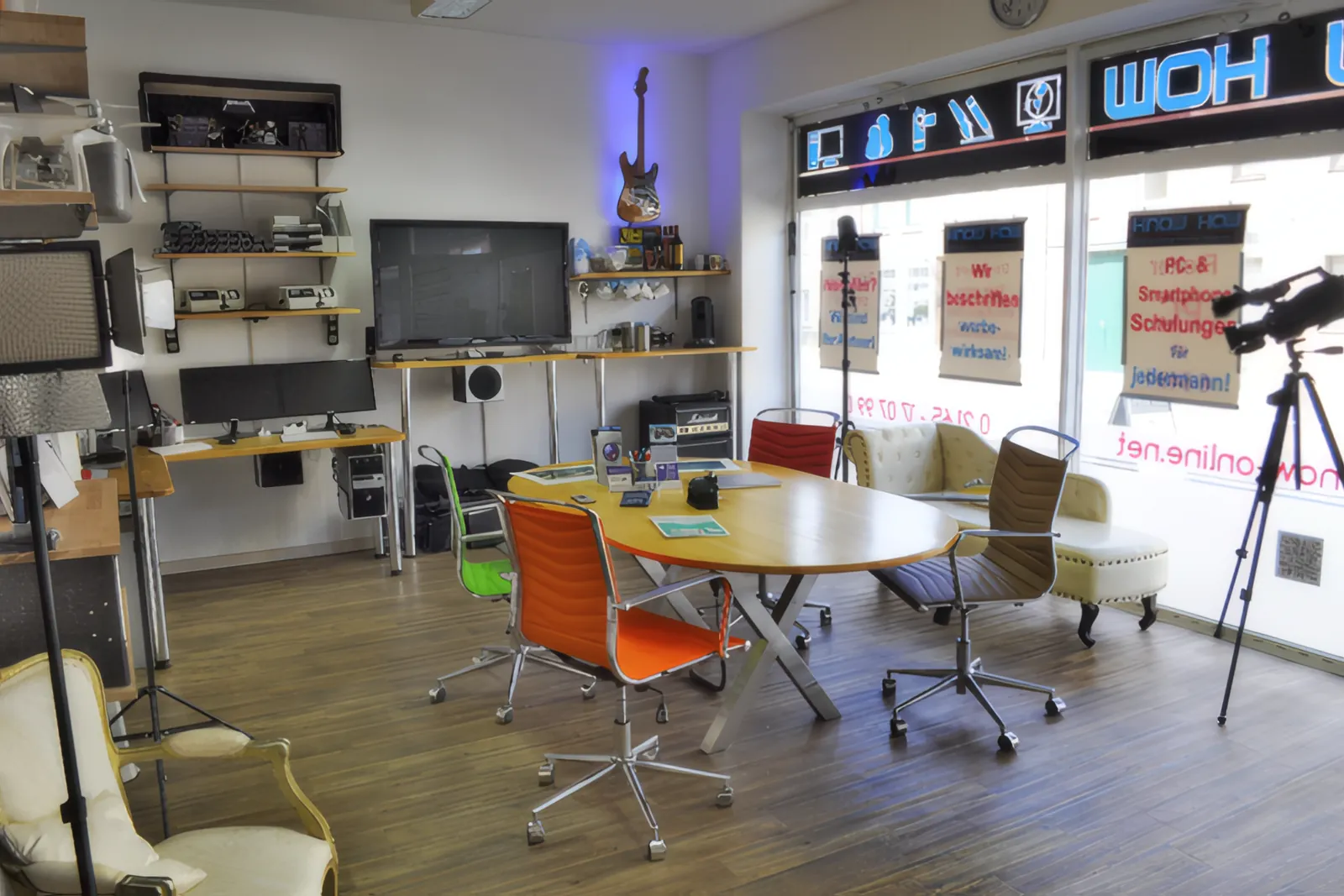 Der Vorraum der Agentur KNOW HOW: ovaler Tisch mit bunten Stühlen, Bildschirm an der Wand, Kamera auf dem Stativ am Schaufenster.