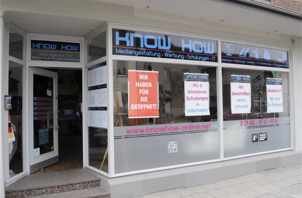 Die Agentur KNOW HOW von außen an der Odenkirchener Straße in Jüchen: Schaufenster mit blauem KNOW-HOW-Schriftzug, Plakaten zu Schulungen und Beschriftung, offene Eingangstür.